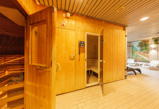 Ingang van een houten sauna in een ontspanningsruimte met ligstoelen en een muurschildering op de achtergrond.
