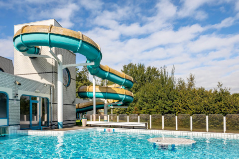 Piscina al aire libre con un gran tobogán acuático en espiral junto a un centro de ocio bajo cielo azul.