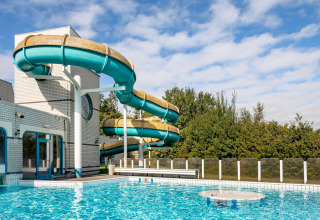 Piscina all'aperto con un grande scivolo d'acqua tortuoso accanto a un centro ricreativo sotto il cielo azzurro.