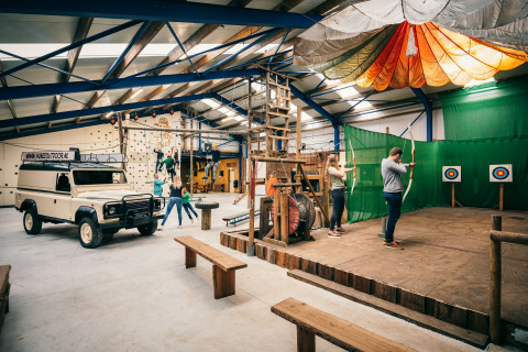 Indoor avonturenpark met boogschieten, klimwand, touwwerk en terreinwagen onder een tentzeil.