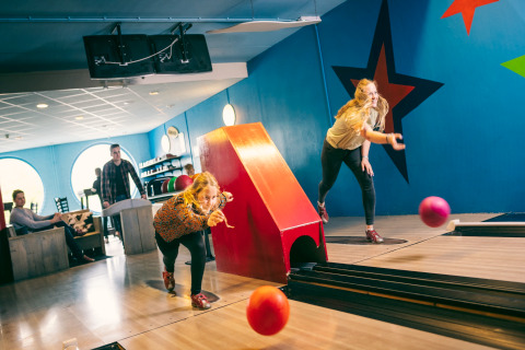 Twee vrouwen bowlen in een kleurrijke bowlinghal terwijl anderen toekijken en ontspannen op de achtergrond.