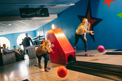 Due donne giocano a bowling in una sala colorata, mentre altre persone guardano e si rilassano vicino.