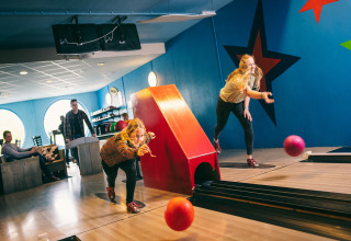 To kvinder bowler i en farverig bowlinghal, mens folk ser til i baggrunden under afslappede forhold.