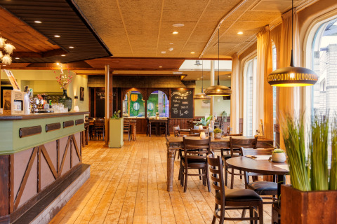 Binnenzicht van een sfeervol restaurant met houten vloer, houten meubilair en grote ramen met gordijnen.