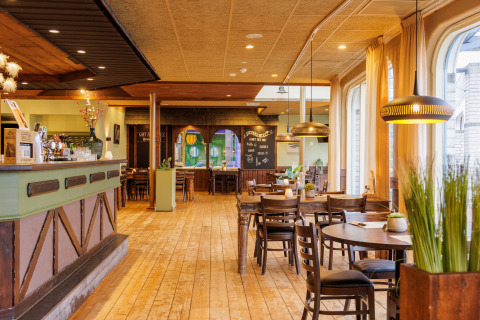 Vista interna di un ristorante accogliente con pavimento in legno, tavoli di legno e grandi finestre con tende.