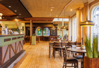 Vista interior de un restaurante acogedor con suelo de madera, mesas de madera y grandes ventanas con cortinas.