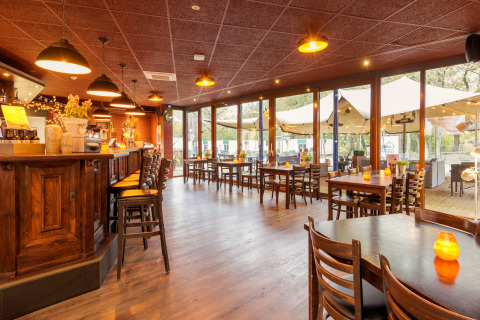 Gezellige restaurantzaal met houten meubilair en bar, zicht op safaritent buiten door de ramen.
