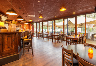 Gezellige restaurantzaal met houten meubilair en bar, zicht op safaritent buiten door de ramen.