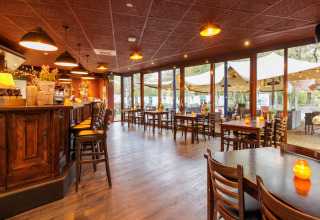 Cozy restaurant interior with wooden tables and bar, large windows facing safari tent setup outside.