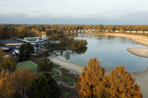 Vue d’un lac, plage de sable, arbres d’automne et bâtiments proches d’une tente safari.