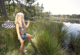 Meisje met stok en zakje verkent het gras bij een meer, vlak bij een safaritent in de natuur.
