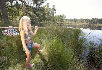 Pige med rygsæk på pind udforsker grønt område nær sø, tæt på safaritelte i naturen.