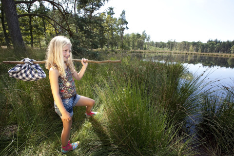 Bambina con bastone e fagotto esplora la riva erbosa di un lago vicino a una tenda safari.