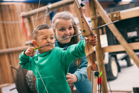 Child learning archery with an adult’s guidance inside a safari tent, both dressed in green.