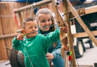 Bambino impara il tiro con l'arco guidato da un adulto in una tenda safari, entrambi in verde.