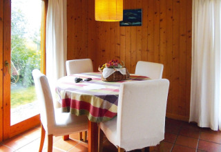Eethoek in bungalow met ronde tafel, vier witte stoelen, kleurrijk tafelkleed en rieten mand als decoratie.