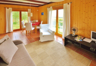 Houten woonkamer in bungalow met grote ramen, eethoek, bank, fauteuil en tv in Portugal.