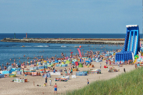 Drukke strand met veel zwemmers, kleurrijke windschermen en een grote blauwe opblaasglijbaan aan zee.
