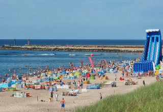 Strand med mange mennesker, farverige telte og stor blå oppustelig rutsjebane ved havet i sommervejr.