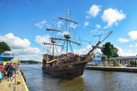 Una nave storica a vela con turisti naviga davanti a un vivace porto sotto un cielo blu e nuvoloso.