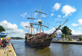 Un barco de vela histórico lleno de turistas navega en un puerto animado bajo un cielo azul despejado.