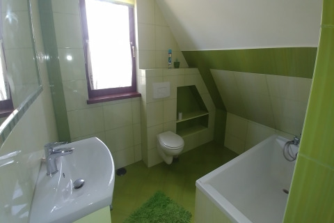Salle de bain lumineuse verte avec baignoire, lavabo moderne, fenêtre et toilettes murales sous plafond mansardé.