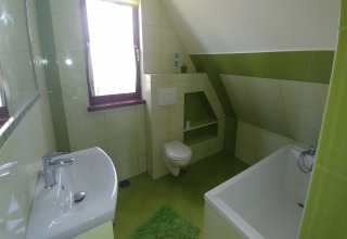 Salle de bain lumineuse verte avec baignoire, lavabo moderne, fenêtre et toilettes murales sous plafond mansardé.