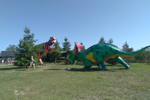Large green and red dinosaur statues in a grassy area with trees and a child running in the foreground.