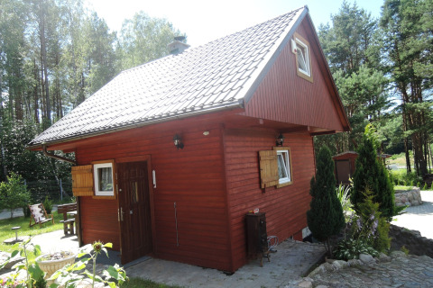 Rød træhytte med hvide vinduer og skråt tag, omgivet af træer i en skovklædt ferieby i Wiselka.