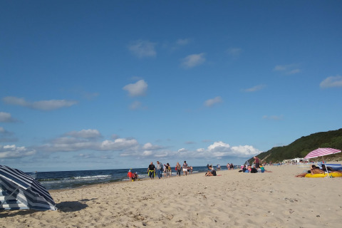 Mensen ontspannen op een strand bij een glamping-accommodatie, met parasols en groene duinen op de achtergrond.