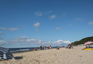 Mensen ontspannen op een strand bij een glamping-accommodatie, met parasols en groene duinen op de achtergrond.