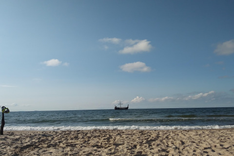 Sandstrand mit Wellen und einem traditionellen Schiff am Horizont, fotografiert bei einer Glamping-Unterkunft.