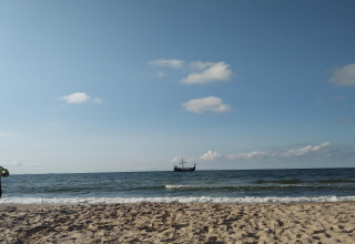 Strand met golven en een historisch schip aan de horizon, genomen bij een glampingverblijf.
