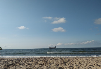 Sandstrand mit Wellen und einem traditionellen Schiff am Horizont, fotografiert bei einer Glamping-Unterkunft.