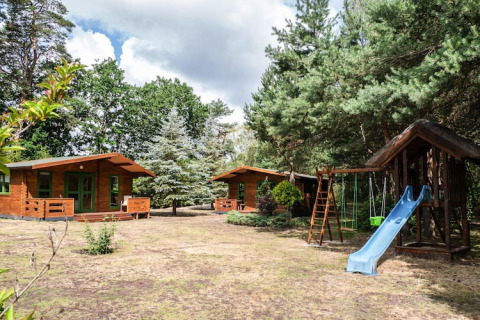 Two wooden holiday homes and a playground with slide and swings set among trees at Kodrabek, Germany.