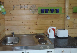 Small wooden kitchen area with sink, stove, electric kettle, toaster, and colorful mugs on shelves.