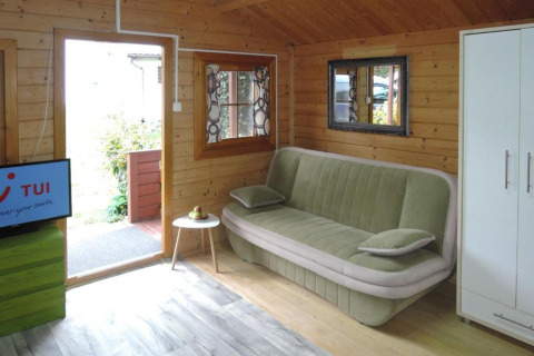 Cozy living area of a wooden cabin with green sofa, TV, small table, mirror, and wardrobe in Germany.