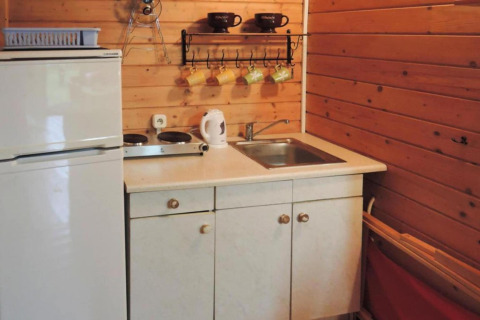 Compact cabin kitchenette with white fridge, sink, kettle, stovetop, and colorful mugs hanging on the wall.