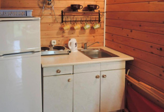 Compact cabin kitchenette with white fridge, sink, kettle, stovetop, and colorful mugs hanging on the wall.