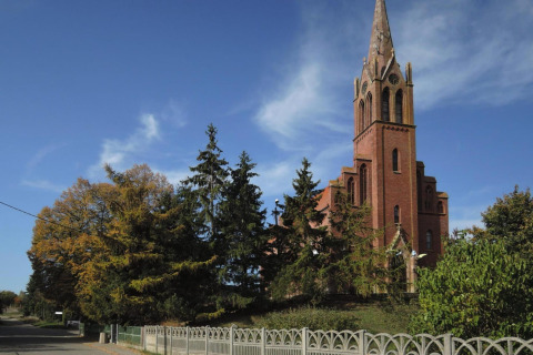 Red brick church with tall spire next to a road, surrounded by green trees and blue sky.