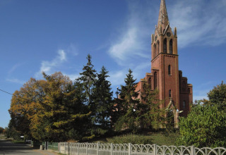 Red brick church with tall spire next to a road, surrounded by green trees and blue sky.