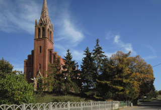 Chiesa in mattoni rossi con alto campanile circondata da alberi e cielo blu vicino a una strada tranquilla in Germania.