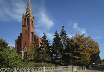 Iglesia de ladrillo rojo con alto campanario rodeada de árboles y cielo azul junto a una calle tranquila en Alemania.