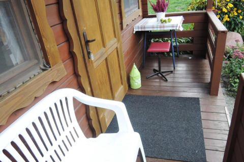 Cabin porch with white plastic bench, yellow door, table, red stool, flowers, and garden in background.