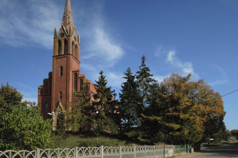 Église en briques rouges à haute flèche, entourée d’arbres et de ciel bleu, vue depuis une route calme.