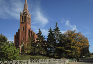 Red-brick church with tall spire and Gothic architecture, surrounded by trees beneath a clear sky.
