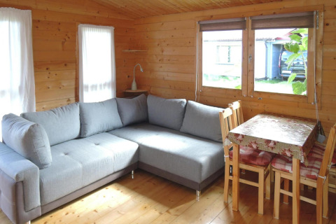 Cozy wooden cabin with a gray sectional sofa, floral tablecloth dining area, and large windows to the outside.