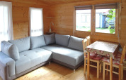 Acogedora cabaña de madera con sofá gris en L, comedor con mantel floral y grandes ventanas al exterior.