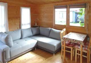 Acogedora cabaña de madera con sofá gris en L, comedor con mantel floral y grandes ventanas al exterior.