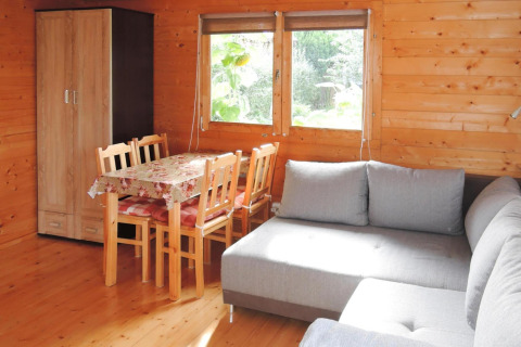 Wooden cabin interior with dining table, four chairs, gray sectional sofa and garden view through windows.